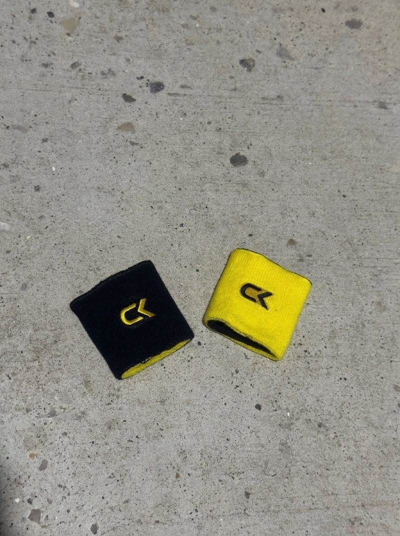 Two Calix reversible wristbands on concrete, one turned black side up and one yellow side up, each with an embroidered CK logo