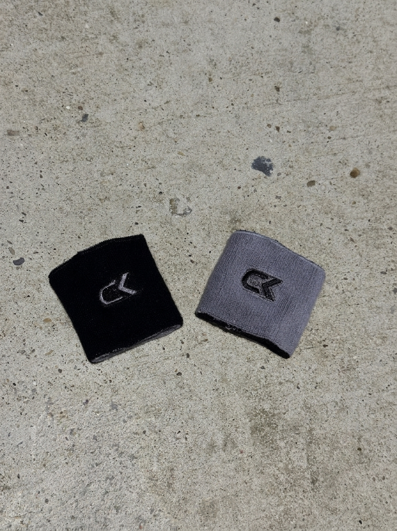 Two Calix reversible wristbands on concrete, one turned black side up and one smoke grey side up, each with an embroidered CK logo