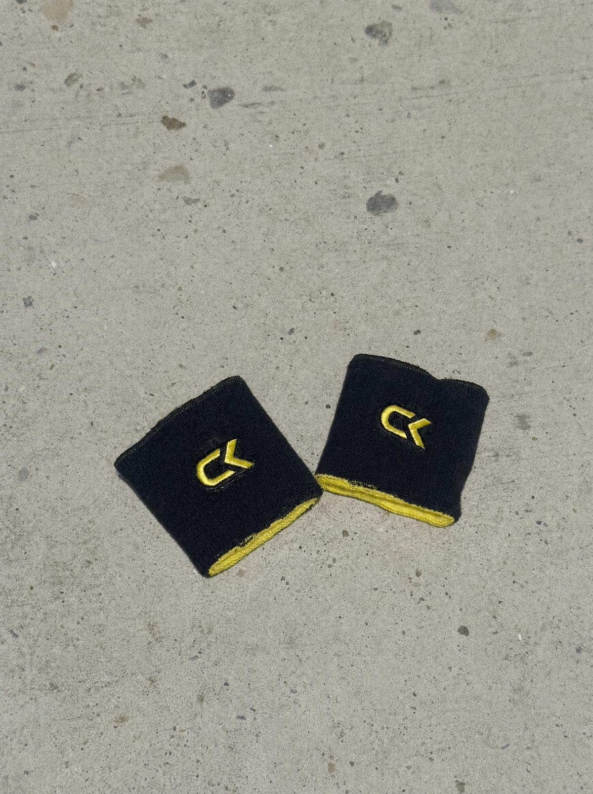 Pair of Calix wristbands worn black side out, yellow terry cotton showing at the trim, embroidered yellow CK logo